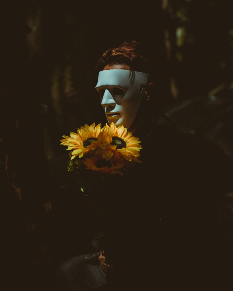 A person wearing a mask holds sunflowers, casting a mysterious and eerie vibe in dim lighting.