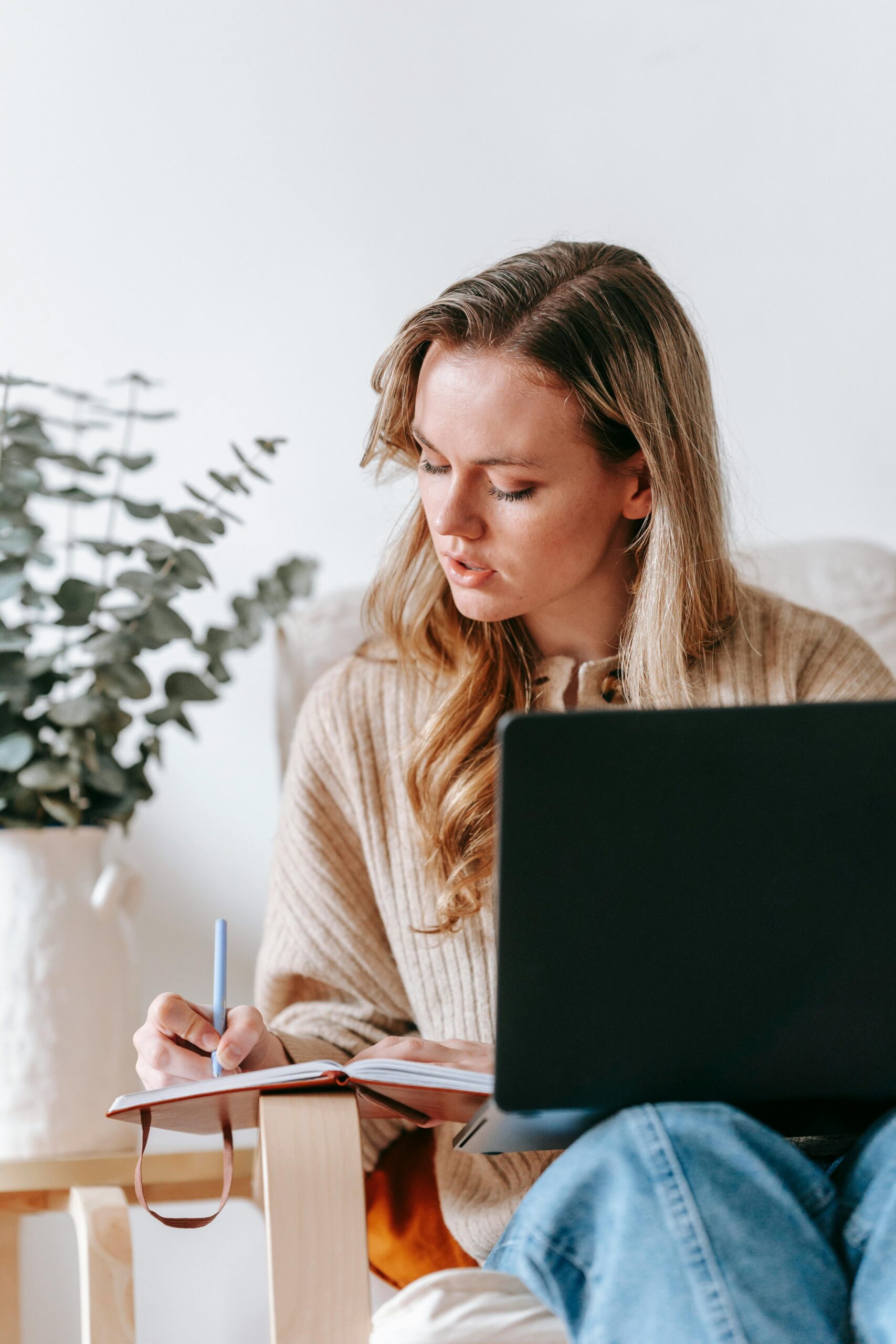 Woman in cozy attire working on a laptop and writing notes in a home setting.